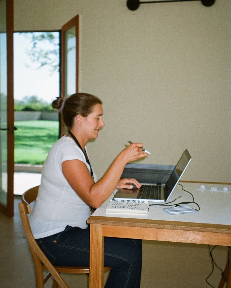 A woman in her 40s who runs a small landscaping company sits at her kitchen table, laptop open beside a paper notebook of client names.